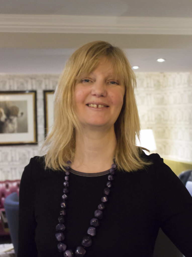 Profile photograph of Susannah. A white woman with shoulder length blonde hair and grey-blue eyes. She is smiling. Susannah is wearing a black dress and a long necklace made of purple, geometrically cut stones.