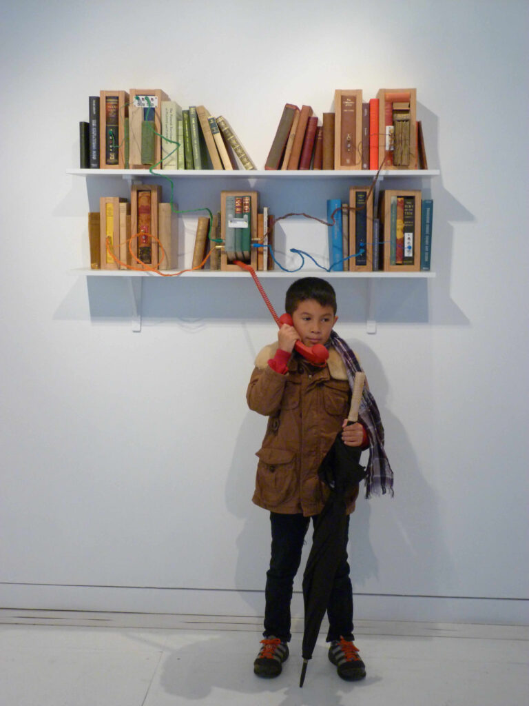'Bibliotech' a book sculpture / installation by Stevie Ronnie. Two 1.2 m shelves, arranged vertically on the white wall of a gallery, are filled with books that have been organised by colour. In among the books are a number of framed sculptures made with recycled books. These framed sculptures are connected together with colourful hand-braided audio cables. In the middle of the bottom shelf there is a famed book sculpture that acts as a cradle for a red telephone handset through which an infinite poem is playing. A young boy with dark skin is holding an umbrella and listening attentively to the poem.