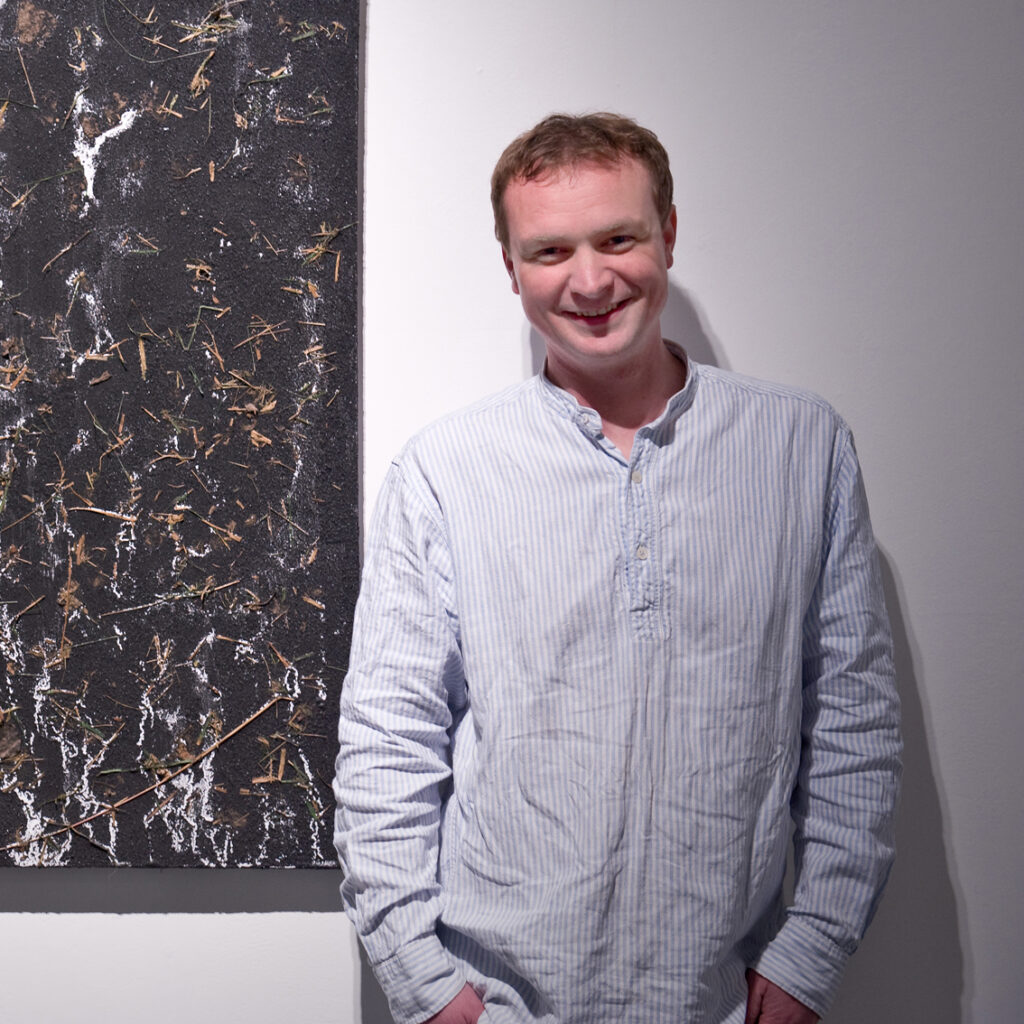 Profile image of Stevie, an average height white male, dressed in a collarless, blue and white striped shirt. He has short brown hair and is smiling, standing casually with his hands in his pockets. He is standing in a gallery, in front of one of his visual artworks - a 1m x 1m piece that was made by melting ice on top of a canvas covered in coal dust.